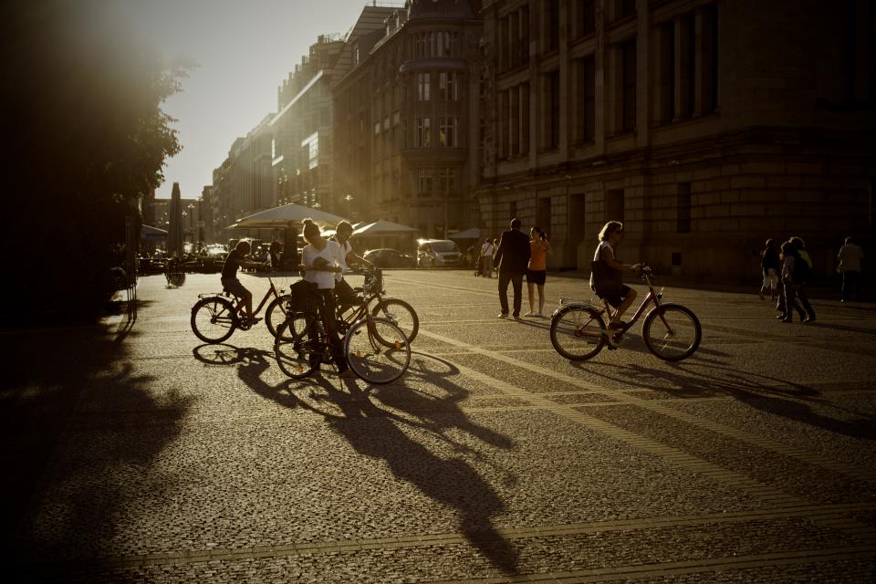 Das Bild zeigt eine sonnendurchflutete Stadtszene mit Menschen, die zu Fuß oder mit Fahrrädern unterwegs sind. Die Schatten der Personen und Fahrräder sind lang und auffällig auf dem gepflasterten Untergrund sichtbar.