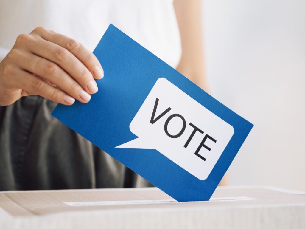 Eine Hand legt einen blauen Wahlzettel mit der Aufschrift "VOTE" in eine Wahlurne.