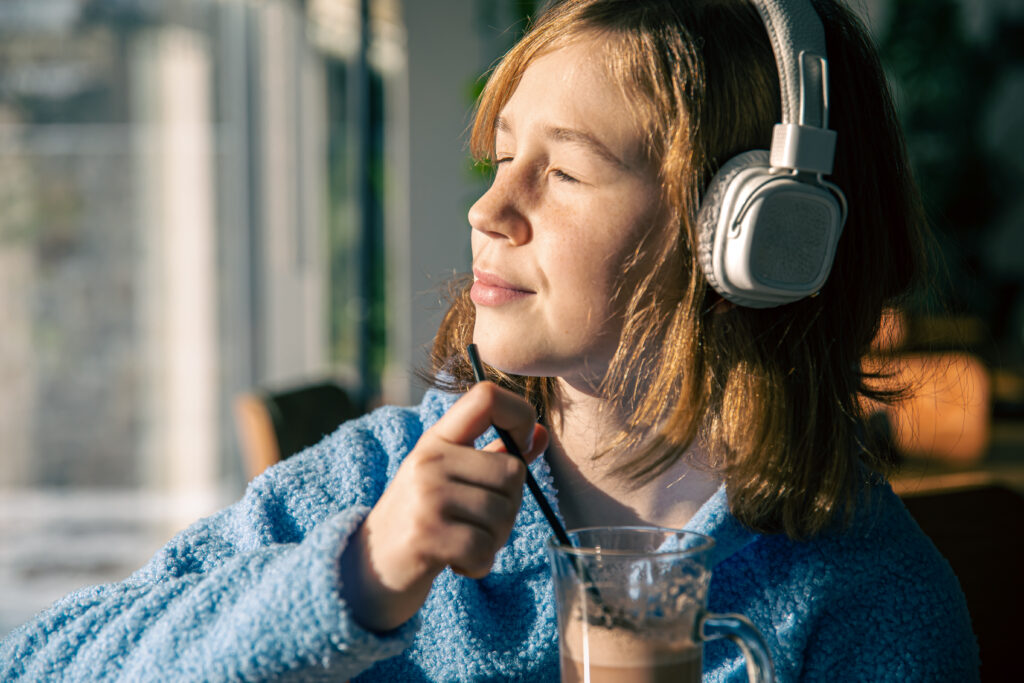 A young women is sitting near the window, listening to music in headphones and drinks cocoa through the tube in sunny weather.
